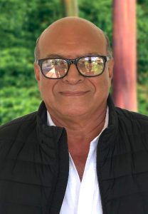 Portrait of a man wearing glasses and a black jacket, standing outdoors with a green background.